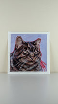 A square art print featuring a painted tabby cat portrait with a light blue background and red accents, displayed against a beige wall on a light surface.
