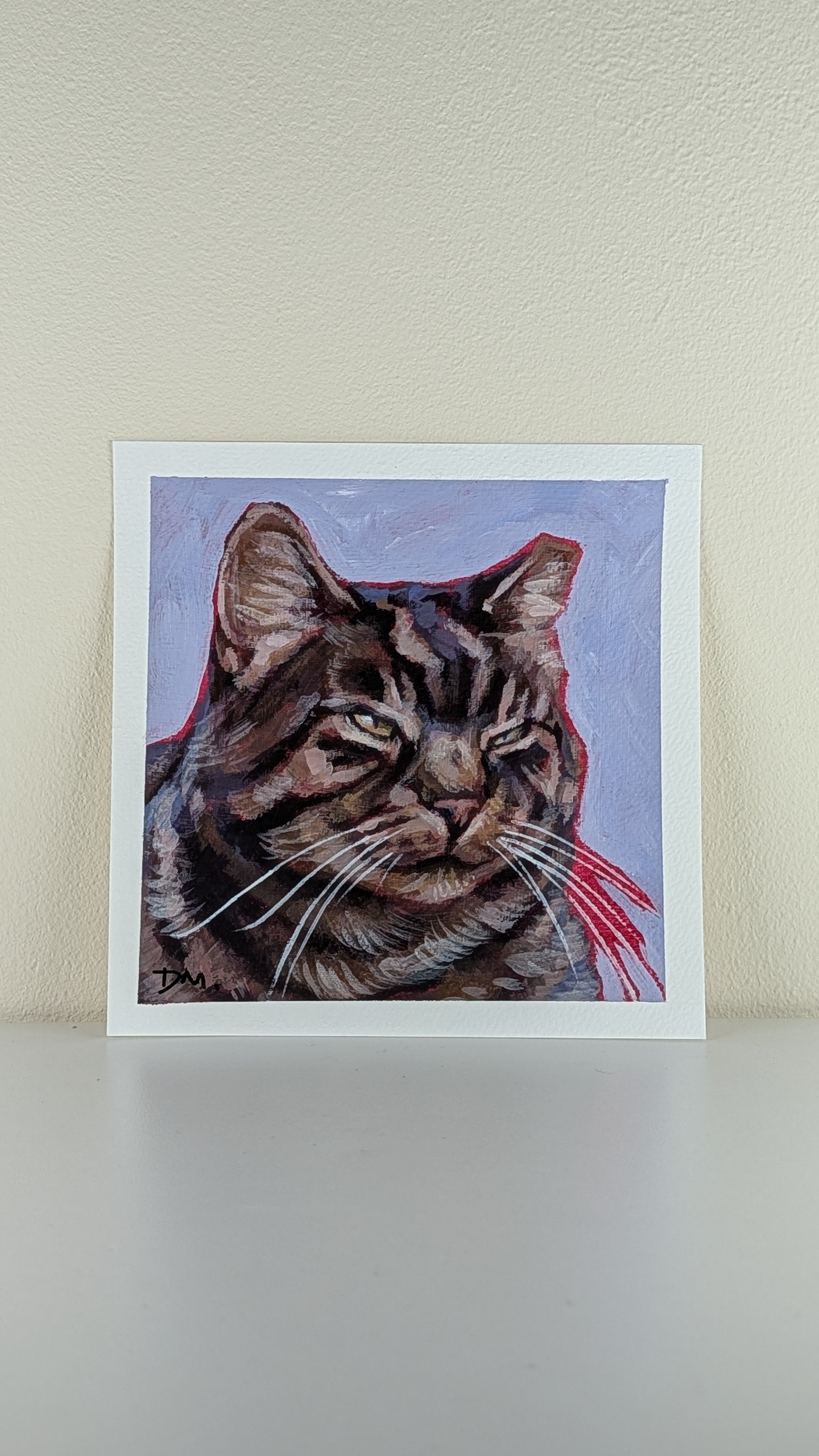 A square art print featuring a painted tabby cat portrait with a light blue background and red accents, displayed against a beige wall on a light surface.